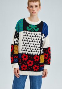 Pull en tricot coloré arborant divers motifs géométriques et floraux en rouge, bleu, vert et blanc, avec un col et des poignets côtelés.