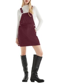 Burgunderfarbene Corduroy-Latzkleid mit verstellbaren Trägern, Fronttaschen-Detail, kombiniert mit knielangen schwarzen Lederstiefeln.