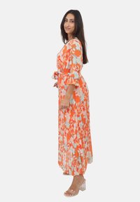 Maxikjole i orange med blomsterprint, der har korte flæseærmer og en bindebånd i taljen, pareret med gennemsigtige block-heel sandaler.