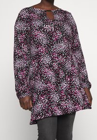 Musta pitkähihainen tunika, jossa on vaaleanpunaisia ja valkoisia satunnaisia pistemäisiä kuvioita, avonaisella kaula-aukolla, yhdistettynä tummanharmaisiin farkkuihin.