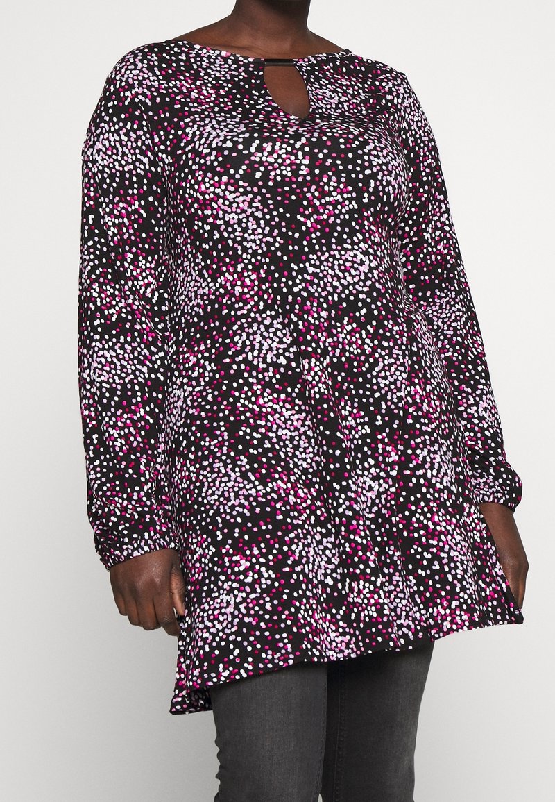 Musta pitkähihainen tunika, jossa on vaaleanpunaisia ja valkoisia satunnaisia pistemäisiä kuvioita, avonaisella kaula-aukolla, yhdistettynä tummanharmaisiin farkkuihin.