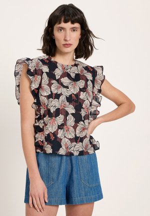 Woman with shoulder-length dark hair wearing a ruffled floral print top and blue denim shorts, posing with hand on hip against a plain background.