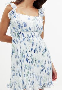 Robe florale en tissu léger, présentant des motifs bleus et verts, un décolleté carré et des bretelles à volants. Texture plissée avec une taille smockée.