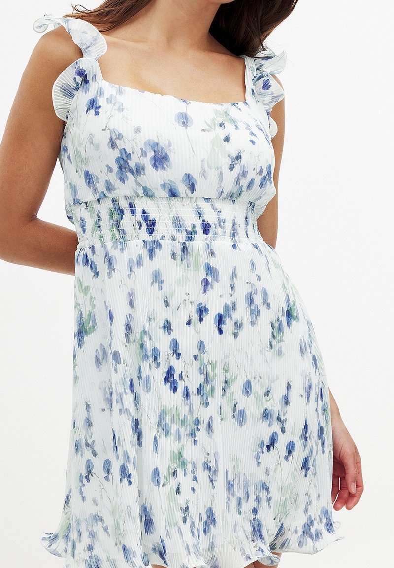 Robe florale en tissu léger, présentant des motifs bleus et verts, un décolleté carré et des bretelles à volants. Texture plissée avec une taille smockée.