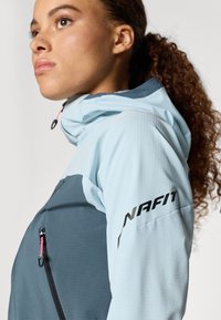 Jauna moteris su garbanotais plaukais, surištais į kuodą, vilkinti šviesiai mėlyną ir tamsiai pilką džemperį su gobtuvu, ant kurio rankovės yra "NAFIT" logotipas, žvelgia į viršų.