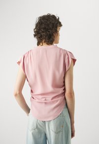 Blusa rosa chiaro con maniche corte a campana, dettaglio arricciato sulla schiena e orlo arrotondato, abbinata a jeans in denim azzurri.