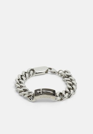 Bracelet en chaîne argentée avec des maillons plats, un accent noir avec un design de croix, et un fermoir en métal gravé du mot "Guess".