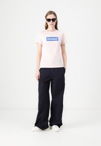 Lys rosa bomull t-skjorte med blå "HUGO" logo, kombinert med løstsittende marineblå bukser og hvite joggesko. Sorte solbriller fullfører stilen.