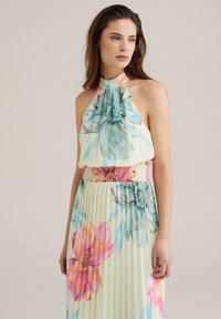 Rochie maxi florală de culoare galben pal, cu guler halter. Are pliseuri, talie ansamblată și imprimeuri cu flori mari în stil acuarelă, în nuanțe de roz și albastru.