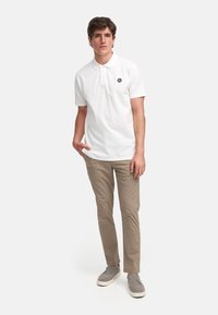 Hvit polo-skjorte med kort krage og svart logo på brystet, kombinert med beige chinos og grå sko. Enkelt, uformelt design.