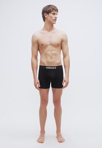 Boxer briefs en coton noir avec taille élastique portant le logo "VERSACE" en blanc. Le modèle est debout, montrant la coupe et le design.