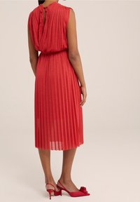 Röd plisserad klänning med rund halsringning och nyckelhålsdetalj bak, tillverkad av lättviktigt tyg. Bärs med matchande röda slingback-klackar.