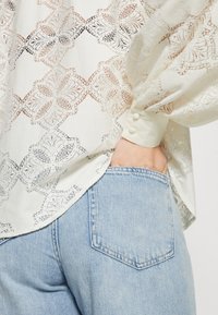 Blouse en dentelle blanche avec motif de papillon et boutons aux poignets, rentrée dans un jean en denim bleu clair, main dans la poche arrière.