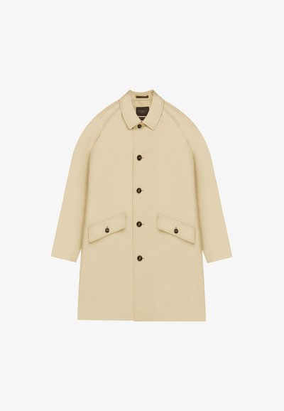 Manteau beige à longueur genou avec un col, de grandes poches avant et des boutons foncés. Présente des manches raglan et une texture de tissu lisse.