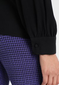 Blusa nera con maniche lunghe a sbuffo e polsini con bottoni, abbinata a pantaloni a quadri viola e neri. Dettagli del tessuto testurizzati visibili.