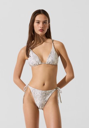 Młoda kobieta w jasnoszarym i białym bikini w wężowy wzór z wiązanymi ramiączkami stoi na tle jednolitego szarego tła.