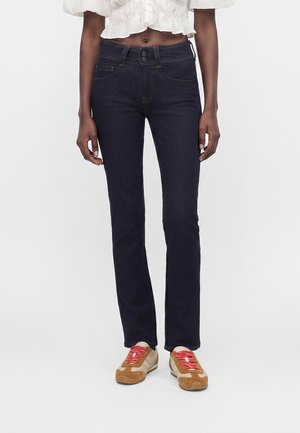 Person wearing dark blue straight-leg jeans, a white cropped blouse, and tan sneakers with red laces, standing against a white background.