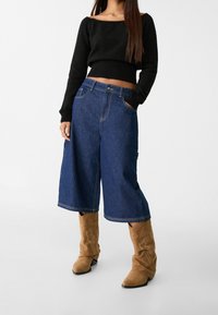 Zwarte cropped trui met een bootnek, gecombineerd met blauwe wijde denim culottes. Tan suède cowboylaarzen maken de look compleet.