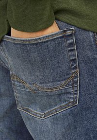 Jeans de mezclilla azul con un bolsillo trasero con costuras en contraste y un ligero desvanecimiento. La cinturilla es parcialmente visible, mostrando una tela verde.