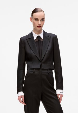 Black cropped tuxedo jacket with satin finish, peak lapel featuring beaded accents, worn over a black shirt and black trousers. White cuffs displayed.