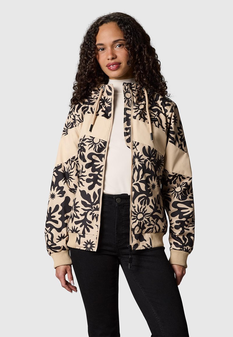 Femme aux cheveux bouclés portant une veste zippée beige et noire à motifs floraux et un pantalon noir, debout devant un fond gris.