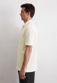 Kurzärmliges, strukturiertes Polohemd in hellem Creme mit einem gerippten Kragen, seitlichen Schlitzen und einer lockeren Passform. Getragen mit dunklen Hosen.