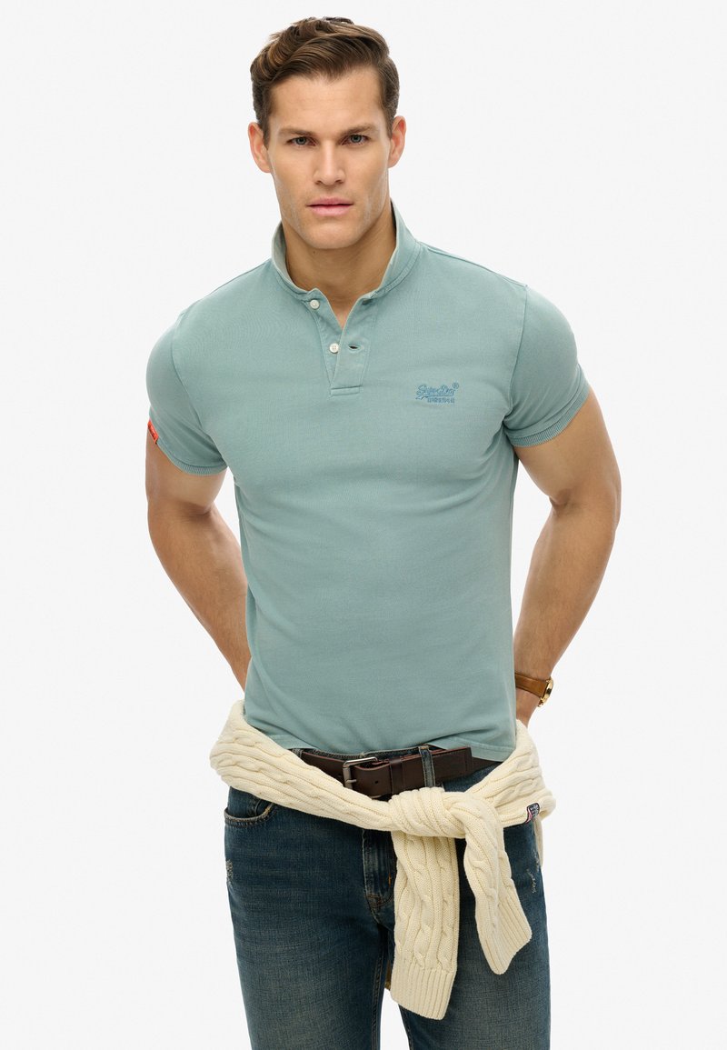 Polo azzurro chiaro con maniche corte, colletto a due bottoni e logo sul petto. Maglione beige legato in vita sopra jeans scuri.