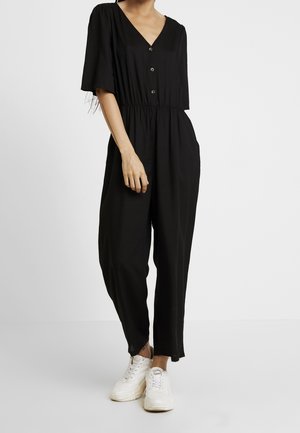 Kvinde iført sort V-hals jumpsuit med knapper, korte ærmer og hvide sneakers, stående foran en ensfarvet lys baggrund.