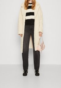 Abrigo esponjoso color crema, suéter a rayas negro y blanco, pantalones gris oscuro y botines negros brillantes. Sosteniendo un pequeño bolso rosa.
