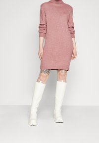 Abito maglione rosa con colletto alto e maniche lunghe, abbinato a stivali in gomma bianchi al ginocchio con suola testurizzata.