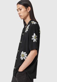 Czarna koszula z krótkim rękawem, ozdobiona białymi wzorami stokrotek, z kieszenią na piersi i okrągłymi guzikami. Miękki materiał o luźnym kroju.