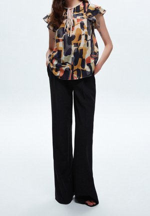 Kobieta w bezrękawnik z abstrakcyjnym wzorem, z falbanami na ramionach i czarnych szerokich spodniach, stojąca z rękami w kieszeniach.