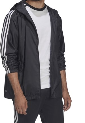 Sort letvægtsjakke med hvide tredobbelte striber på ærmerne, justerbar hætte og elastiske manchetter, båret over en hvid t-shirt med rund hals.