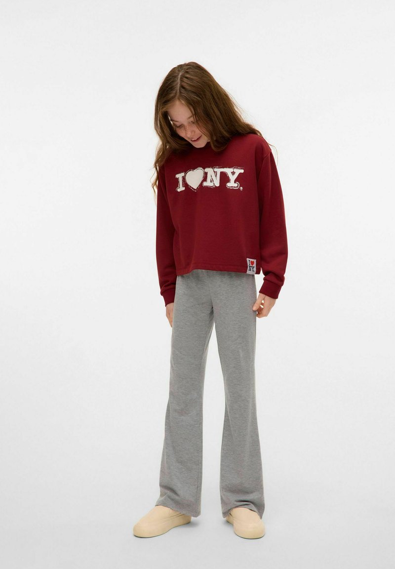 Burgundowa, krótka bluza z nadrukiem "I ❤️ NY"; połączona z jasnoszarymi spodniami dresowymi w kształcie dzwonów i beżowymi butami na slip-on. Gładki materiał, luźny krój.