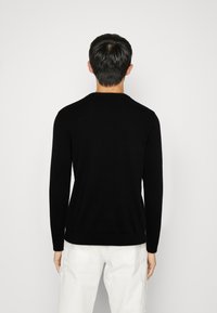 Černý svetr s dlouhým rukávem a kulatým výstřihem, vyrobený z pletené látky. Má úzký střih a žebrované manžety. Model má na sobě světlé kalhoty.