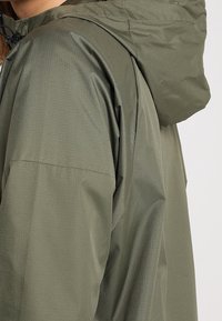 Chaqueta impermeable verde oliva con un acabado texturizado, que cuenta con una capucha ajustable y un corte holgado. Bolsillo en la costura lateral visible en la manga.