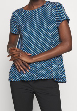 Kobieta ubrana w niebieską, wzorzystą bluzkę z krótkim rękawem ozdobioną okrągłymi wzorami, w połączeniu z czarnymi spodniami, z delikatnie skrzyżowanymi rękami na wysokości talii.