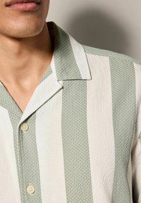 Hellgrünes und weiß gestreiftes Hemd aus strukturiertem Stoff, mit einem Kentkragen und Knopfverschluss. Auffällige Knopfdetails sichtbar.