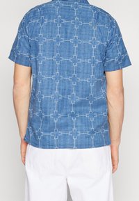 Modrá košile s krátkým rukávem z texturované látky, zdobená bílým geometrickým vzorem. Rovný spodní lem a detaily na zadní části.