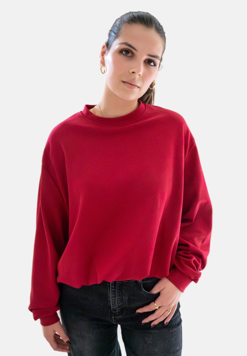Jeune femme avec des cheveux bruns en queue de cheval, portant un t-shirt rouge à manches longues et un jean noir, se tenant avec une main dans la poche contre un fond blanc.