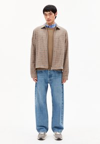 Jonge man in een beige geruite zip-up jack, bruine trui, blauwe shirt, losse blauwe jeans en grijze sneakers, staand tegen een witte achtergrond.