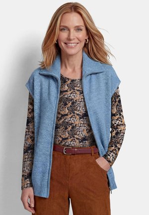Frau mit schulterlangen blonden Haaren, die eine blaue ärmellose Strickweste, ein gemustertes langärmeliges Oberteil und braune Hose mit Gürtel trägt, lächelt.