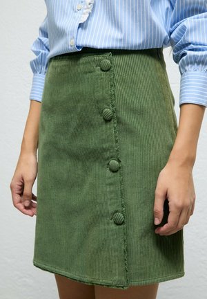 Primer plano de falda de pana verde con cuatro botones forrados de tela, llevada con blusa azul de rayas con mangas puñadas.