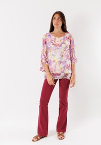 Blusa fluida e floreale con sfumature di viola, rosa e giallo, abbinata a pantaloni rossi aderenti e sandali marroni. Vestibilità rilassata, tessuto leggero.