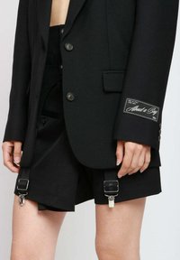 Schwarzes, tailliertes Blazerjackett mit Knöpfen und Branding am Ärmel, kombiniert mit hochtaillierten schwarzen Shorts mit Clip-Verschlüssen.