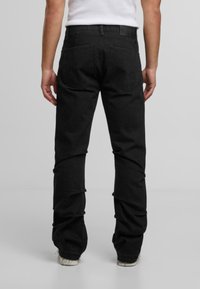 Pohľad zozadu na osobu v čiernych rovných džínsoch, bielych teniskách a zasunutom bielom bezrukávovom tričku na jednofarebnom pozadí.