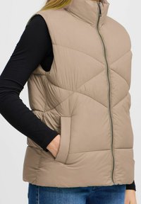 Beige pufferväst med hög krage, dragkedja fram och diagonalt quiltat mönster. Har sidofickor och är överlagd över en svart topp.