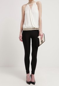 Haut cache-cœur blanc sans manches avec un décolleté en V et une bordure métallique, associé à des leggings noirs et des talons hauts noirs, tenant une pochette noire et blanche.