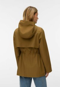 Femme vue de dos portant une veste de pluie à capuche marron avec taille cintrée, associée à un jean bleu, sur un fond blanc uni.