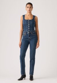 Γυναίκα που στέκεται φορώντας αμάνικο τοπ από σκούρο μπλε τζιν με κουμπιά, τζιν ψηλόμεσο σετ που ταιριάζουν, χρυσούς κρίκους στα αυτιά και μαύρα μποτάκια μέχρι τον αστράγαλο.